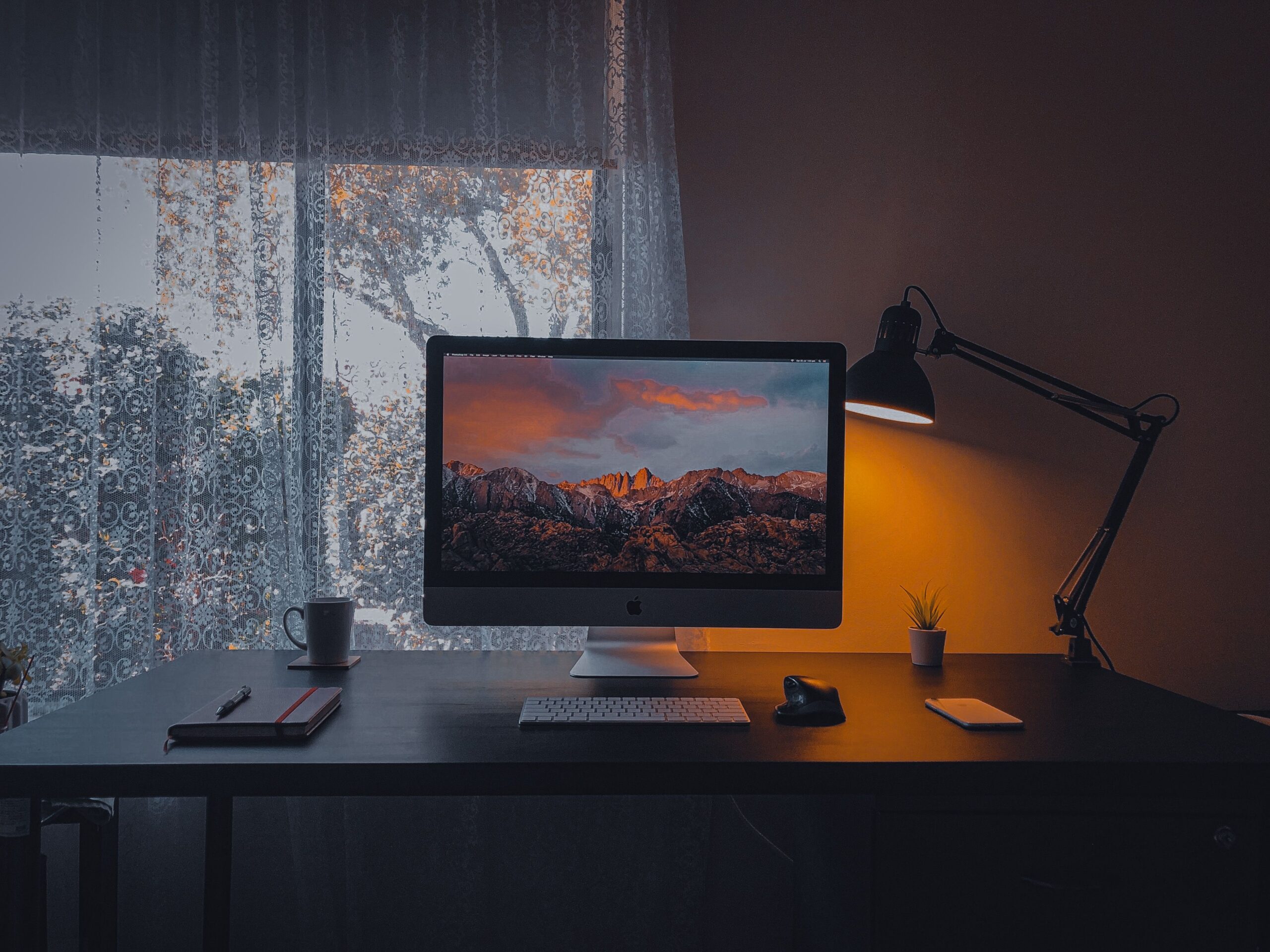 Modern computer with mountain range on screen and desk lamp composing with smartphone and copybook on table near window in dark cozy room